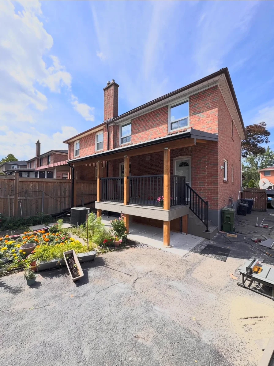 Covered wood deck with railings and steps beside a brick home
