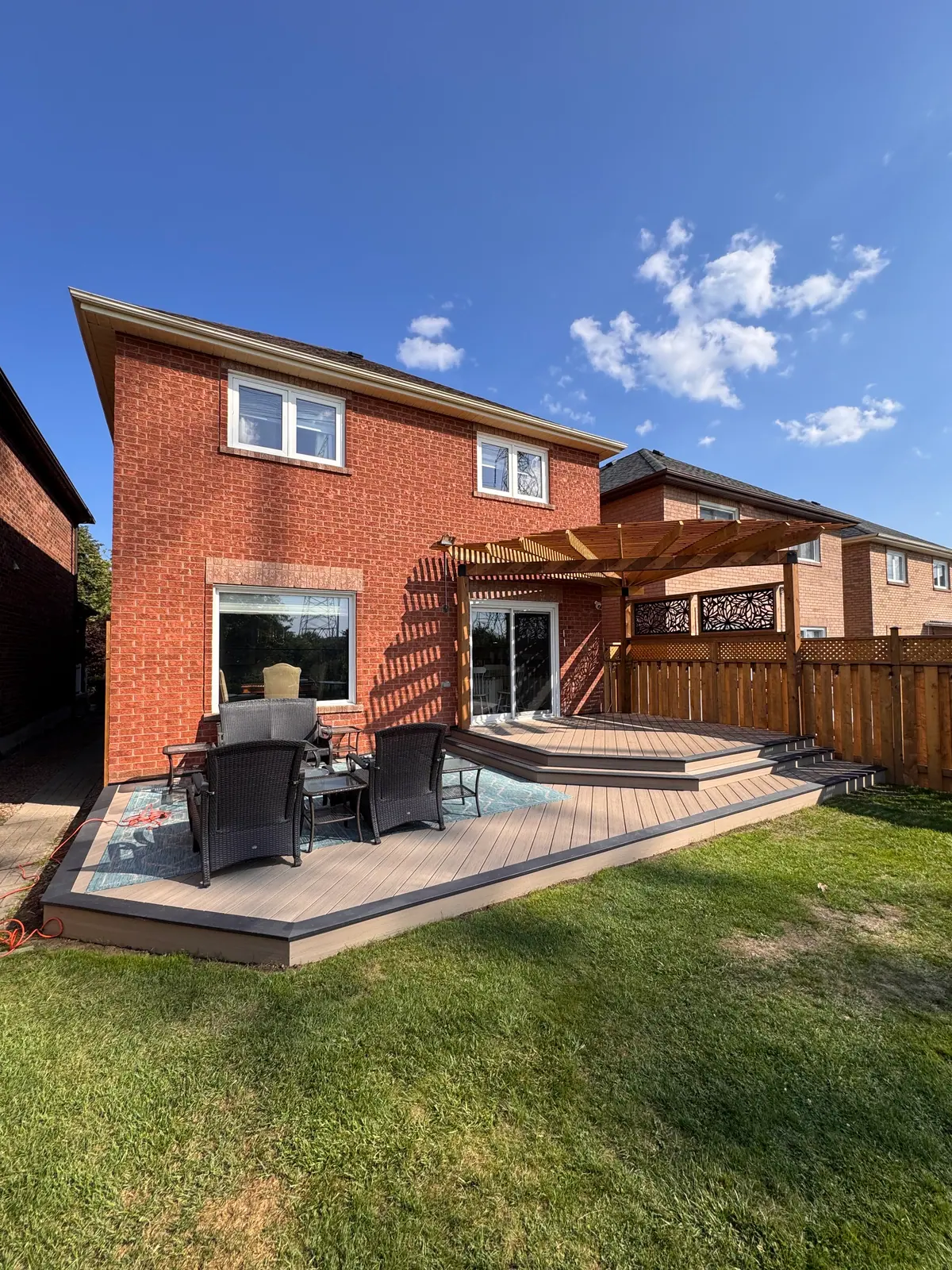 Wide backyard deck with a pergola, steps and outdoor seating beside a brick house