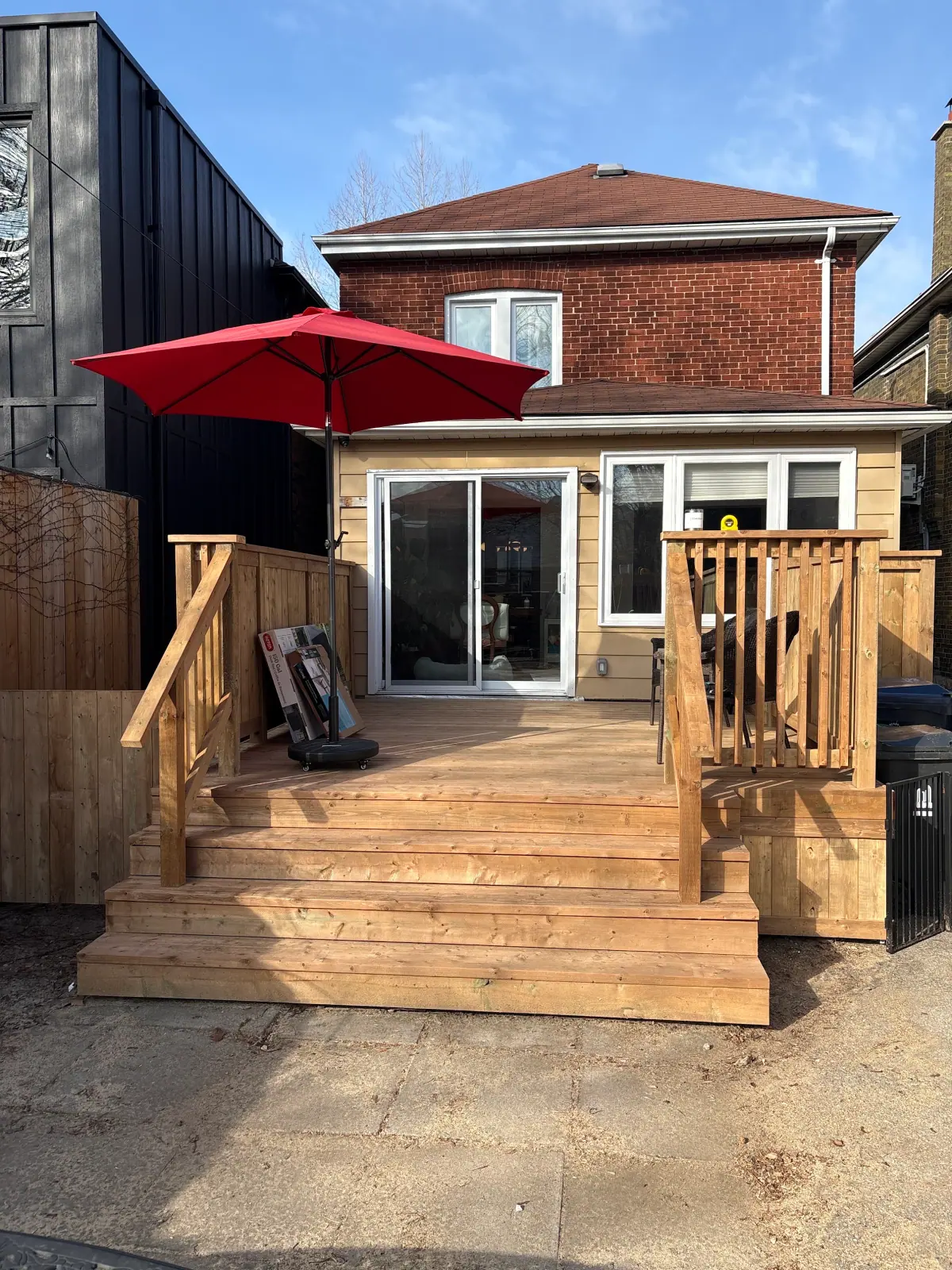 Wooden backyard deck with steps, railings and a red patio umbrella beside a brick home