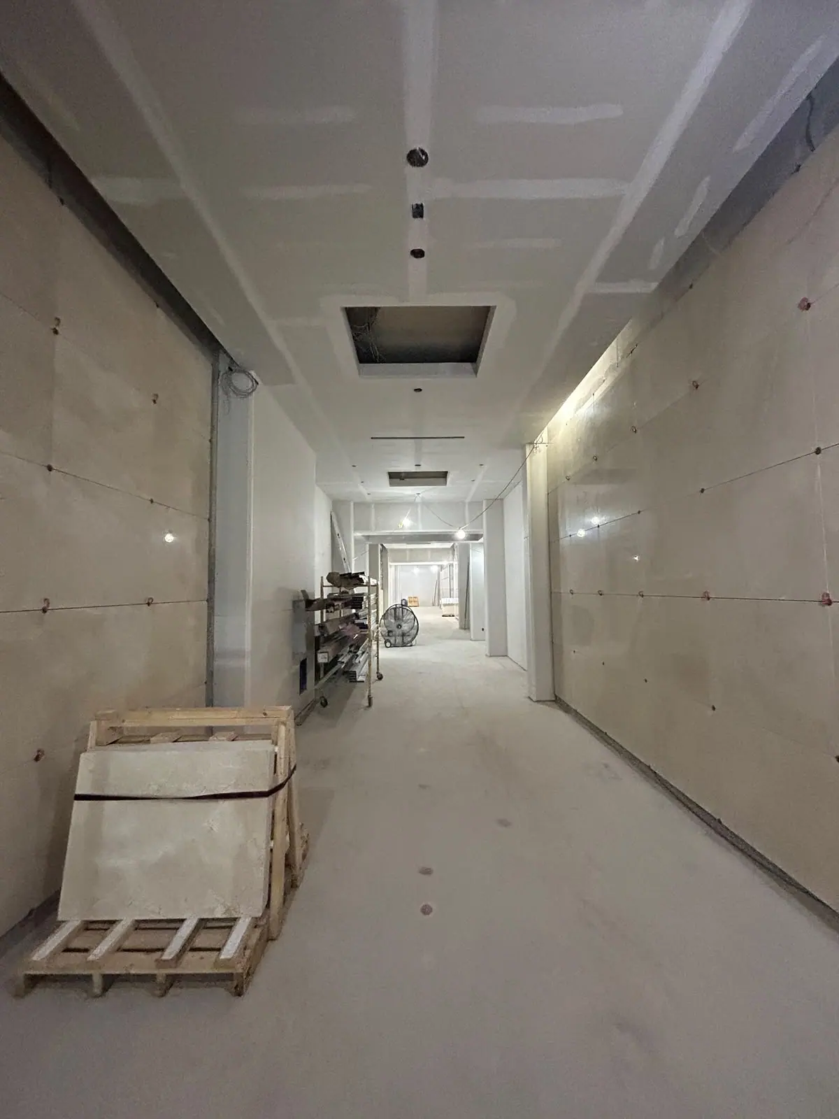Straightened view down an unfinished corridor with drywall panels, taped joints and open ceiling cutouts
